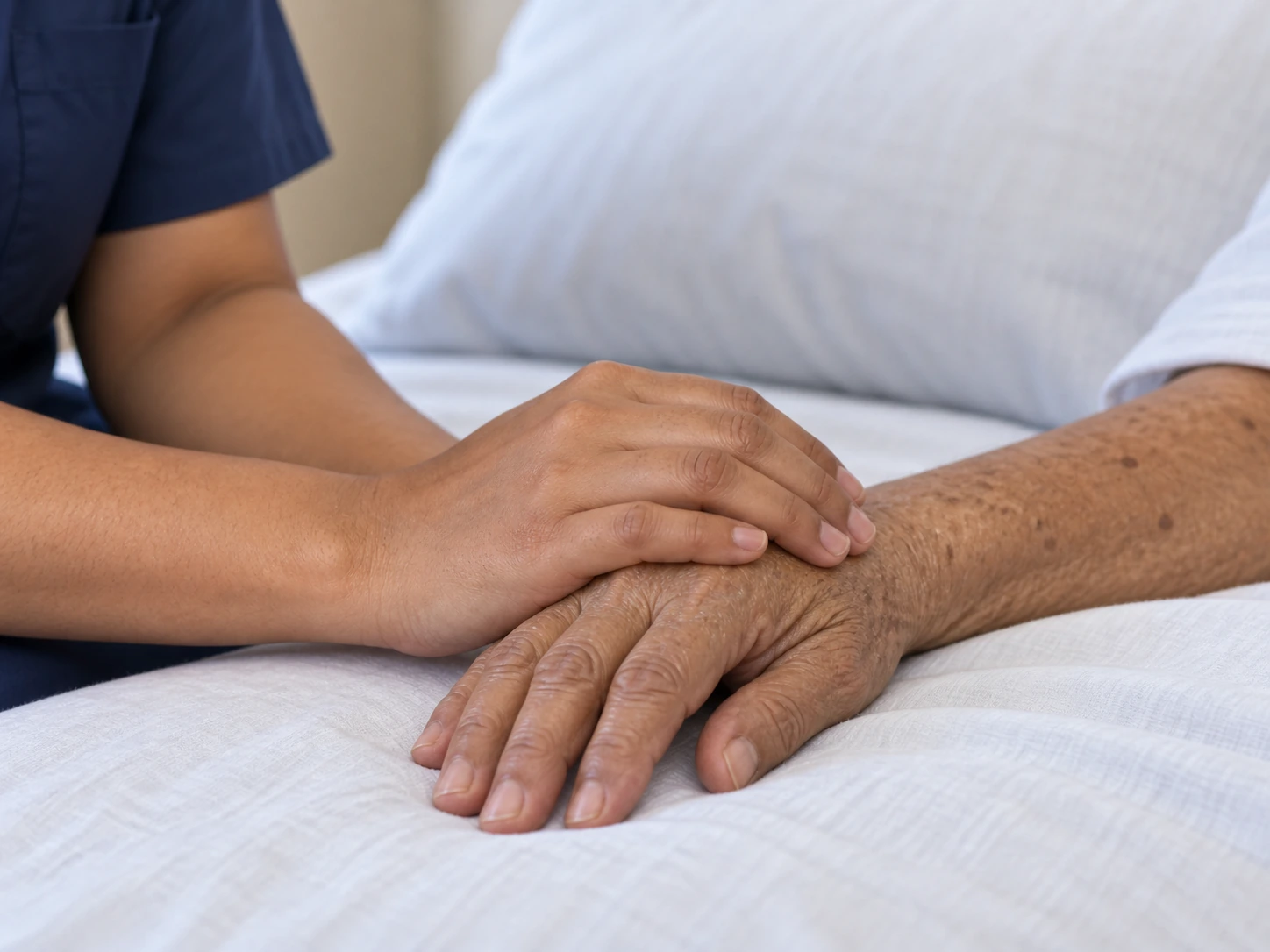 A nurse rests a reassuring hand on an older person’s hand in a home bedroom.